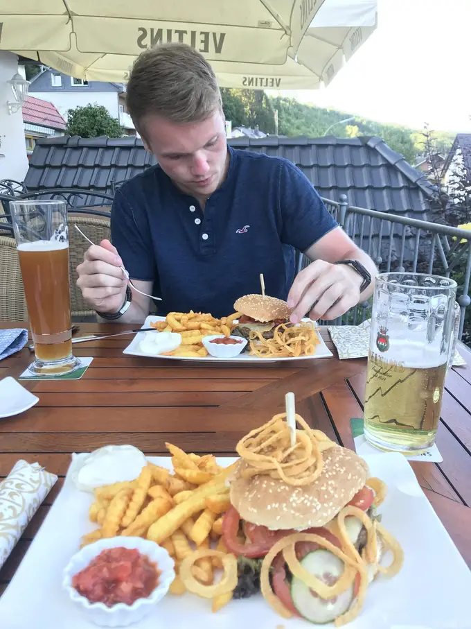 Max isst auf der Terrasse unter einem Sonnenschirm einen Burger mit Pommes, vorne ein zweiter Teller mit Burger, Zwiebelringen und Pommes, daneben ein Weizenbier und ein Bierkrug