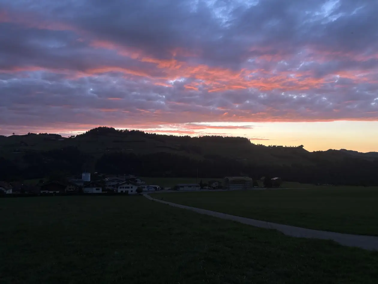 Rosa und violette Wolken über dunklen Hügeln, darunter eine Wiese und ein paar Häuser im Dämmerlicht