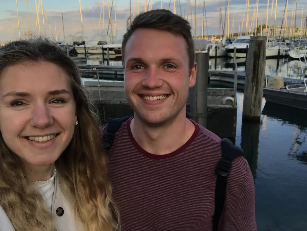 Selfie von Rebecca und Max am Abend an einem Bootssteg, hinter ihnen Segelboote und Masten im letzten Sonnenlicht