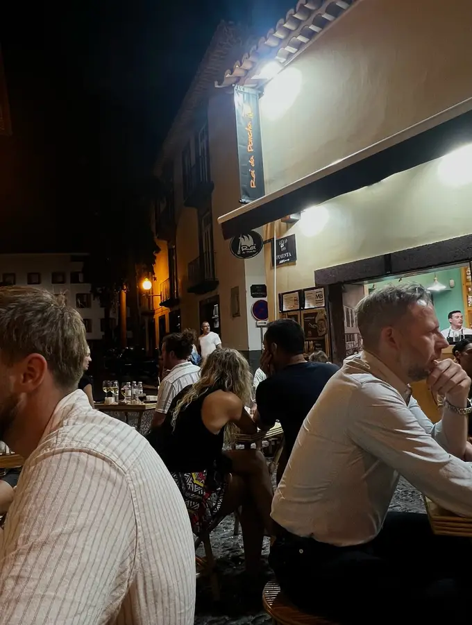 Nächtliche Gassenszene vor einer Bar mit Gästen an Tischen im Freien