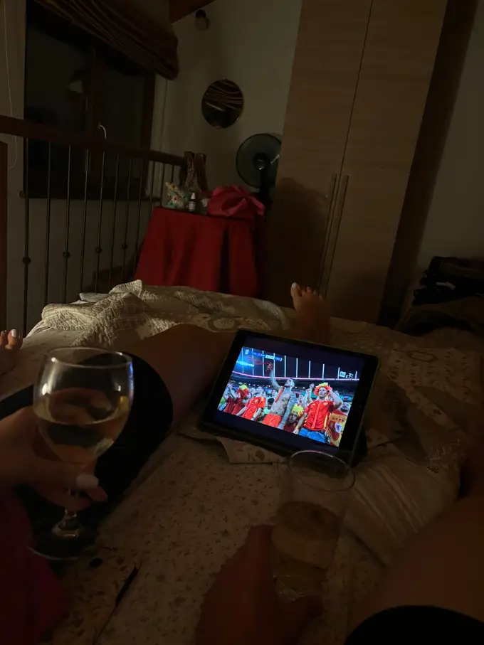 Abends im Bett, auf der Decke ein iPad mit einem Fußballspiel Spanien gegen Frankreich, im Vordergrund zwei Hände mit Weingläsern