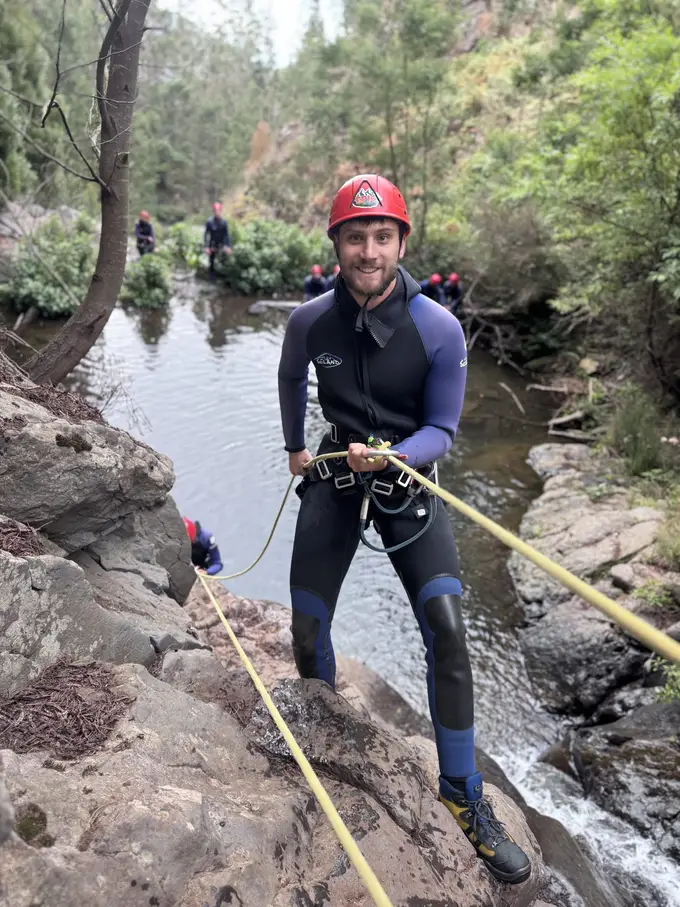 Mann im Neoprenanzug seilt sich an einem Seil eine Felswand neben einem Wasserfall hinab
