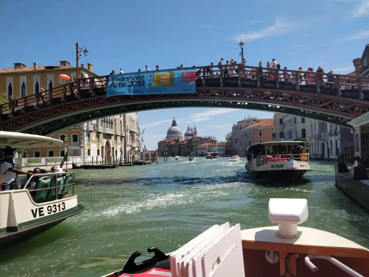 Blick vom Boot auf die hölzerne Accademia-Brücke über dem Canal Grande, darunter ein Vaporetto, in der Ferne die Kuppel der Salute