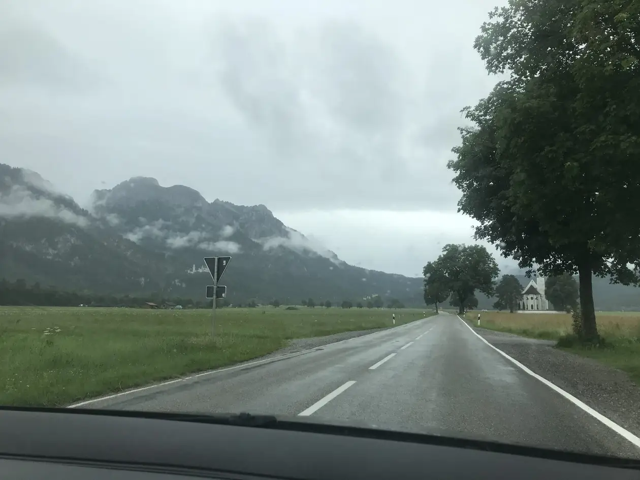 Blick durch die Windschutzscheibe auf eine nasse Landstraße zwischen Wiesen, links Berge im Nebel mit einem kleinen weißen Schloss am Hang, rechts eine weiße Kirche
