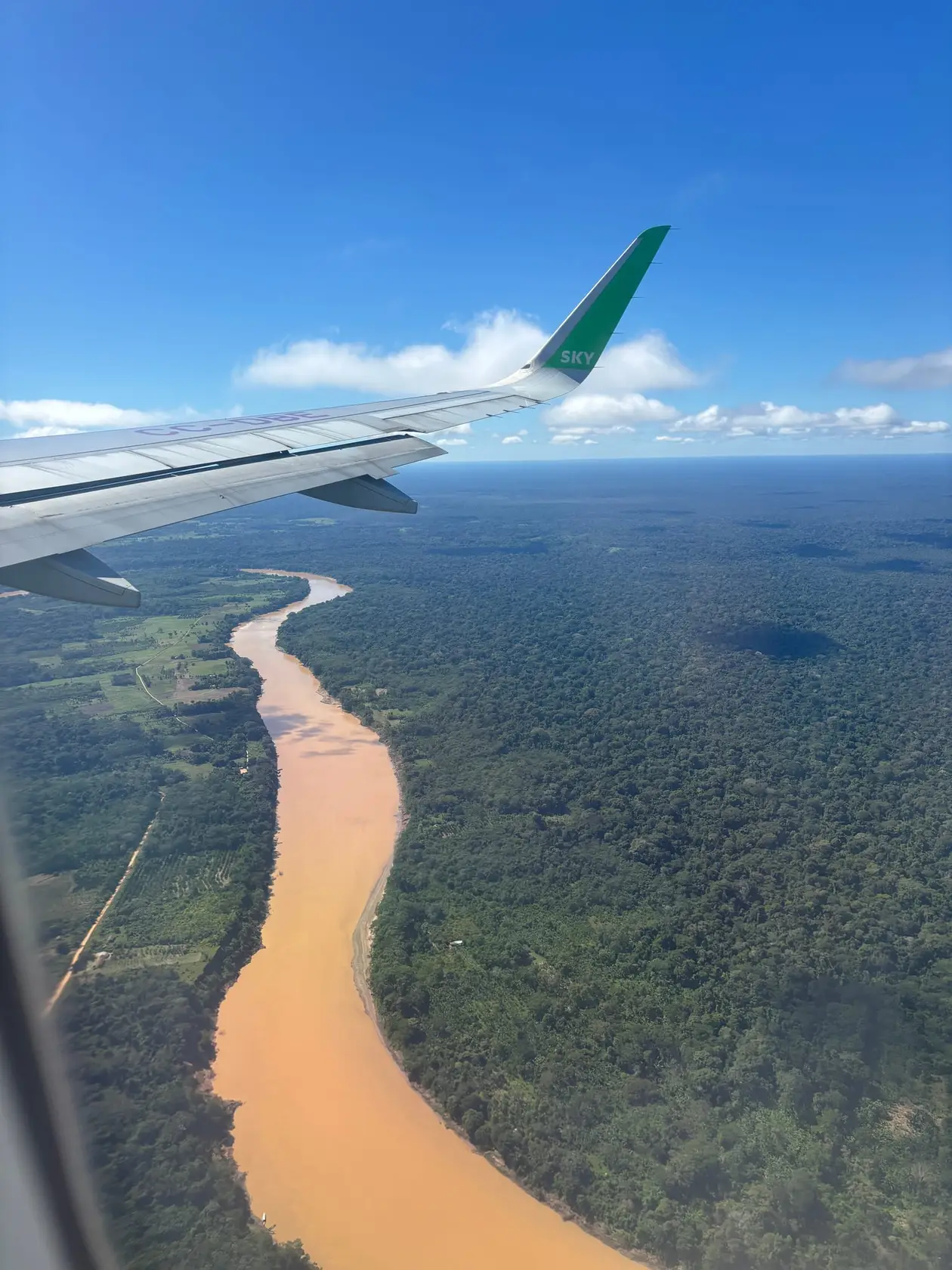 Blick aus dem Flugzeugfenster auf einen breiten, lehmbraunen Fluss, der sich durch dichten grünen Regenwald zieht, oben die grüne Flügelspitze