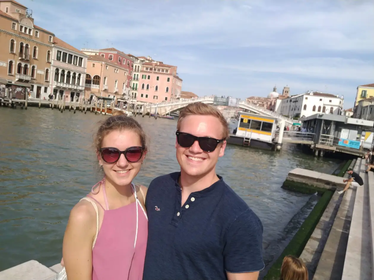 Rebecca und Max mit Sonnenbrillen am Ufer des Canal Grande, hinter ihnen eine steinerne Brücke und eine Vaporetto-Station