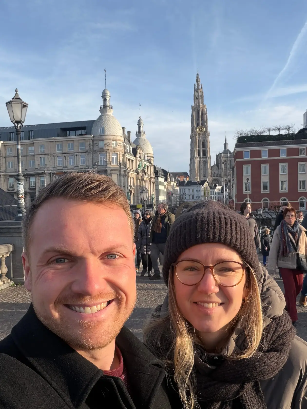 Selfie von Max und Rebecca in der Sonne, hinter ihnen Passanten, ein Kuppelgebäude und ein hoher gotischer Kirchturm