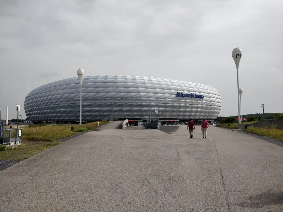 Die Allianz Arena mit ihrer silbrig-weißen Rautenfassade und dem blauen Schriftzug unter bedecktem Himmel, auf dem breiten Vorplatz zwei Männer von hinten