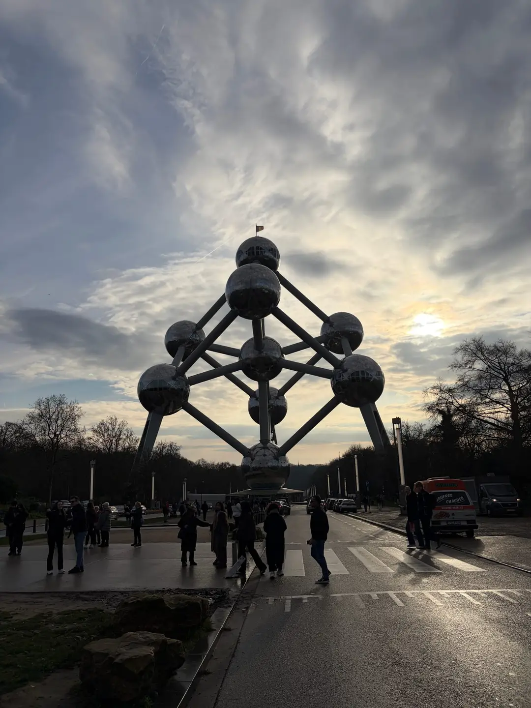 Das Atomium im Gegenlicht als dunkle Silhouette vor bewölktem Himmel, davor Menschen auf dem Vorplatz und ein Waffelwagen