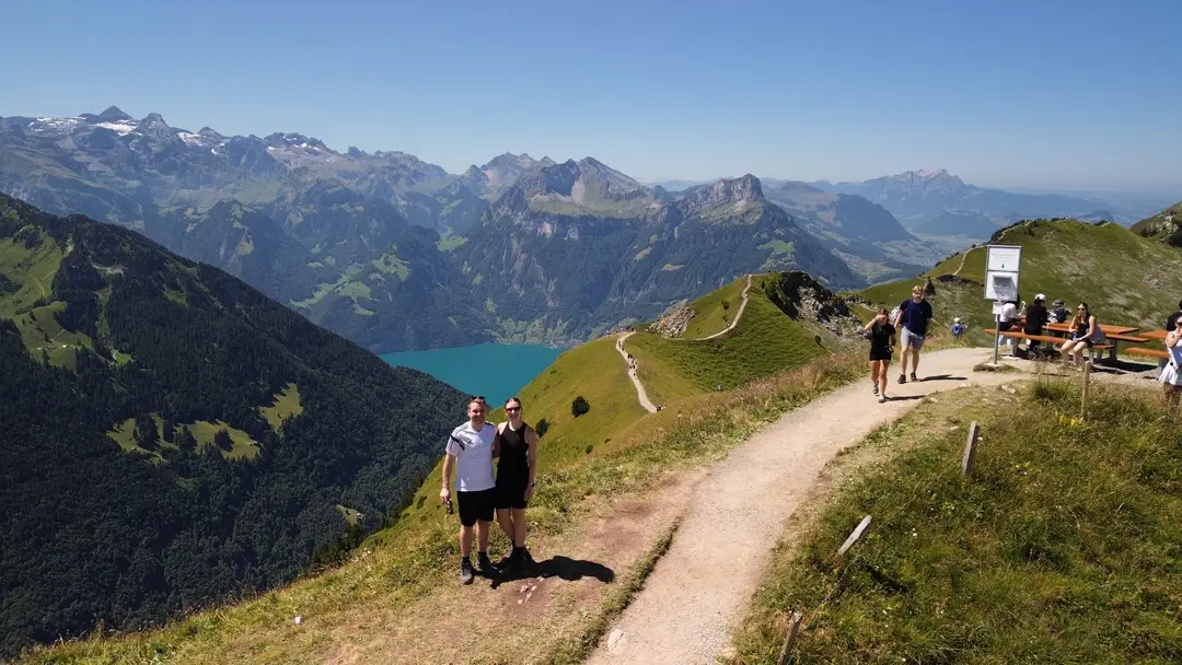 Drohnenfoto: Max und Rebecca stehen auf dem Gratweg, links fällt der Hang zu einem türkisen See ab, dahinter Bergketten