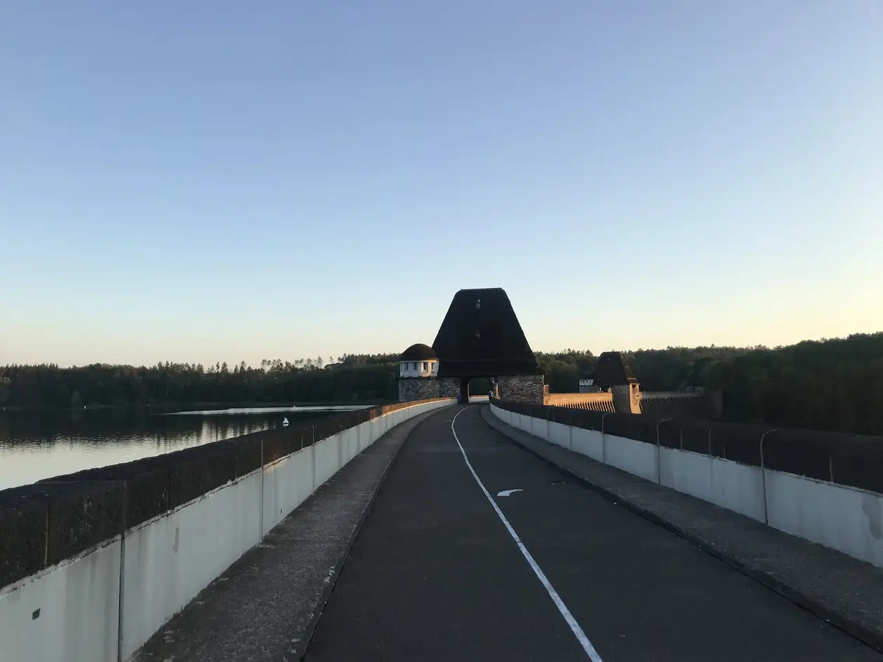 Der Weg auf der Krone der Möhnetalsperre zwischen zwei Brüstungen führt auf einen Turm mit hohem, spitz zulaufendem Dach zu, links liegt der Möhnesee im Abendlicht