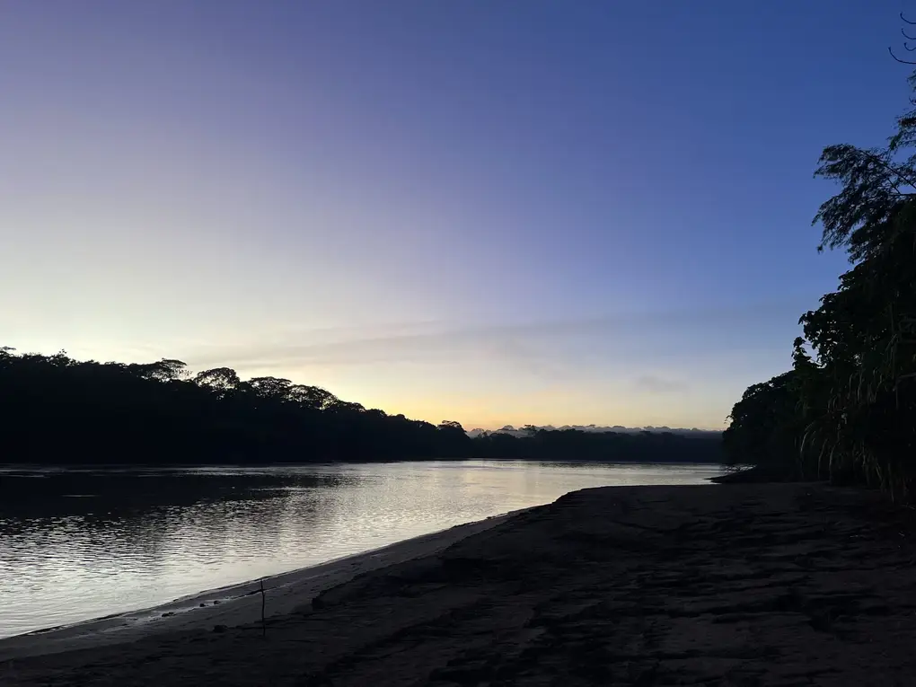 Fluss in der Morgendämmerung, vorne eine dunkle Sandbank, am Horizont ein heller Streifen über dem Wald