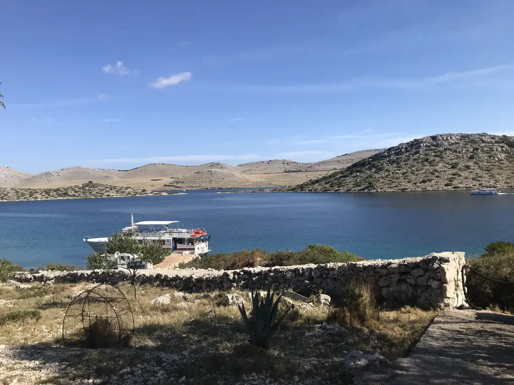 Ein Ausflugsboot mit Sonnendach liegt an einem Anleger in einer Bucht, dahinter kahle Hügel, vorne eine Trockenmauer und eine Agave