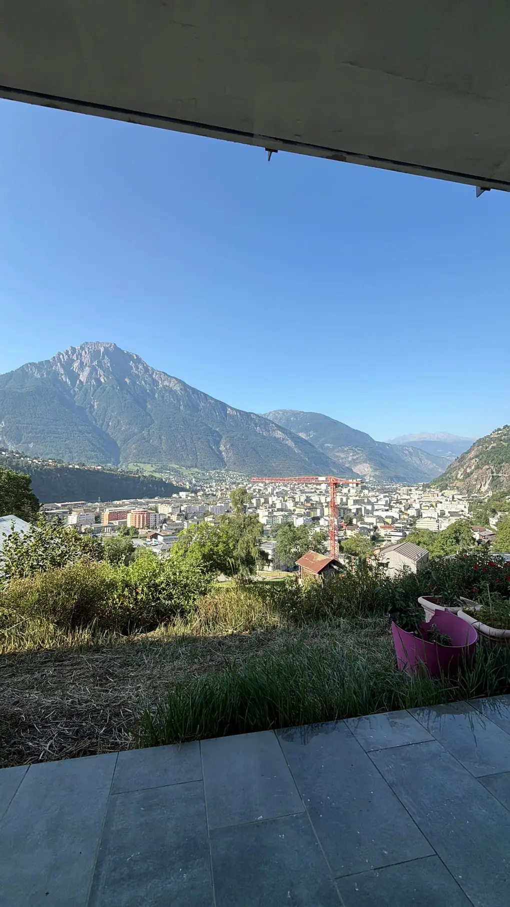 Blick unter dem Terrassendach hervor über Gras und Sträucher auf die Stadt im Tal und einen spitzen Berg unter blauem Himmel