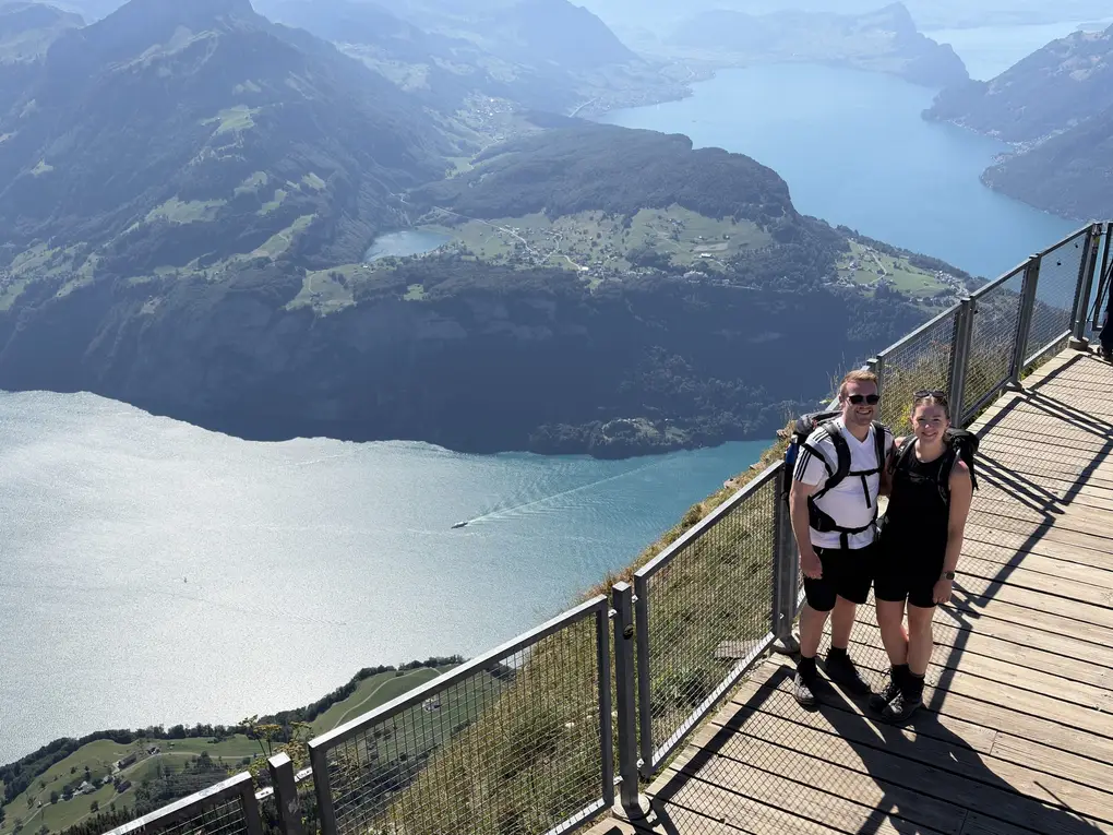 Max und Rebecca auf einer Aussichtsplattform aus Holz mit Geländer, weit darunter ein großer See zwischen bewaldeten Bergen