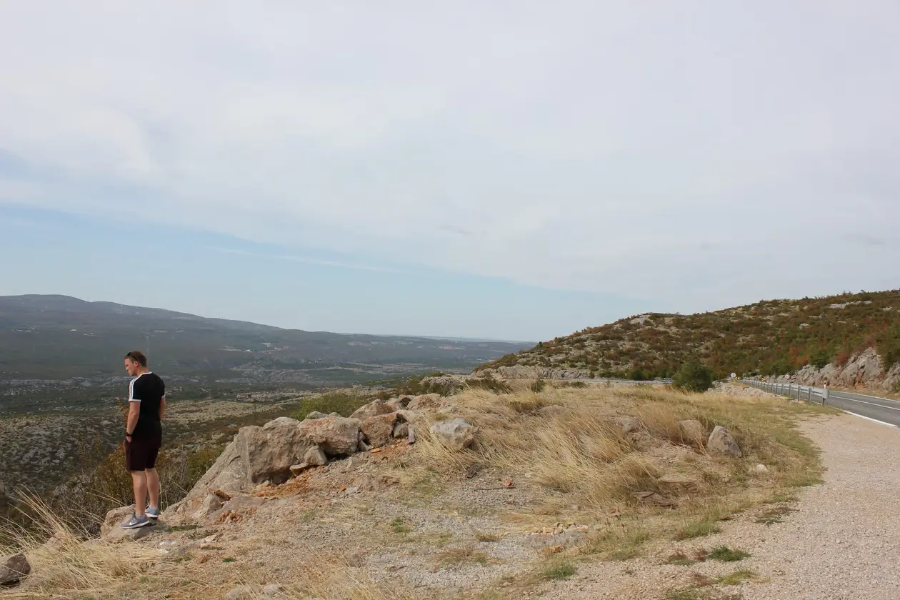 Max steht auf einem Felsen am Rand einer Landstraße und schaut über eine weite, trockene Karstebene, am Horizont Windräder