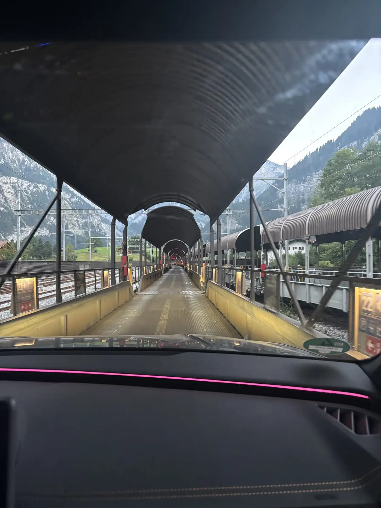 Blick durch die Windschutzscheibe über das Armaturenbrett auf einen überdachten Autozugwaggon, links und rechts Gleise und Berge
