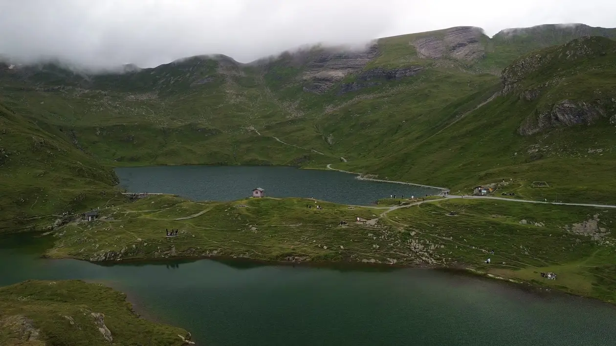 Drohnenfoto über zwei grünlich dunkle Bergseen, dazwischen ein Wiesenrücken mit Wegen und einer kleinen Hütte, oben tief hängende Wolken