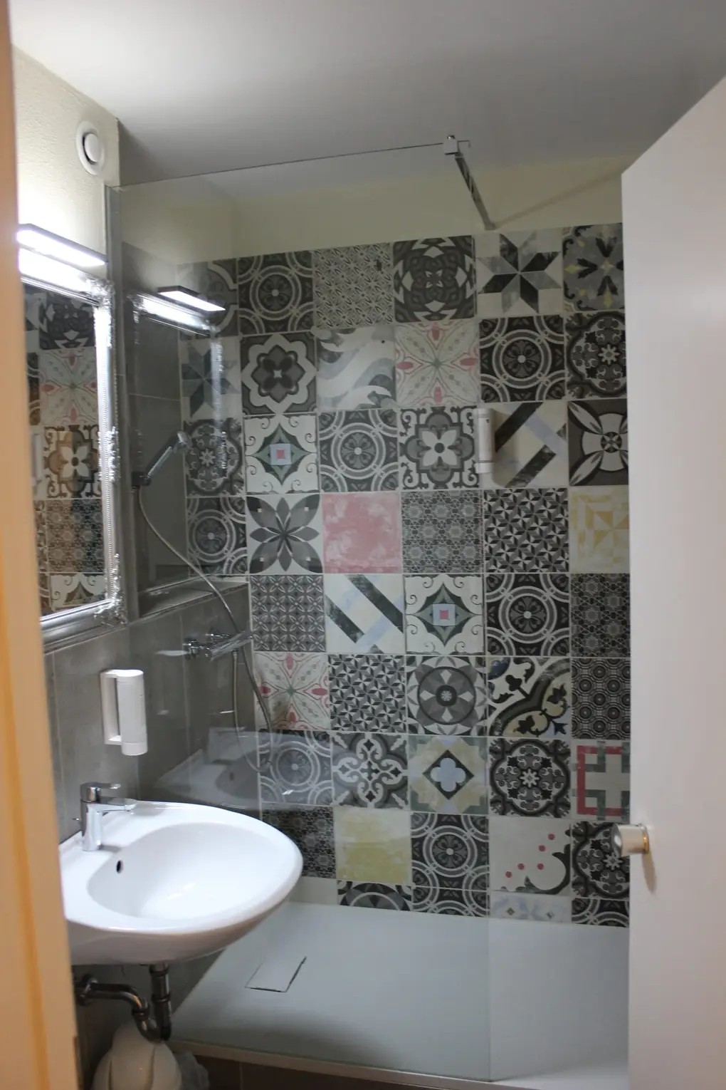 Kleines Bad mit Waschbecken und ebenerdiger Dusche hinter Glas, die Wand bunt gemustert mit Zementfliesen