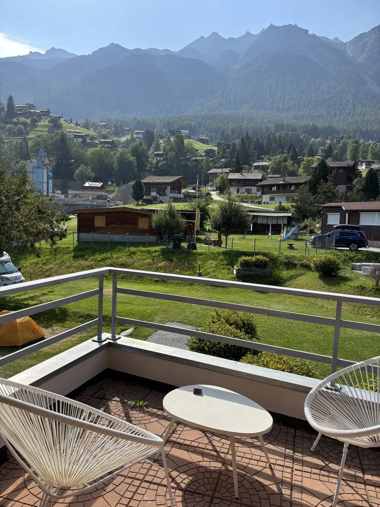 Balkon mit weißem runden Tisch und zwei weißen Schnurstühlen, darunter eine Wiese und Holzhäuser, dahinter bewaldete Hänge und eine Bergkette