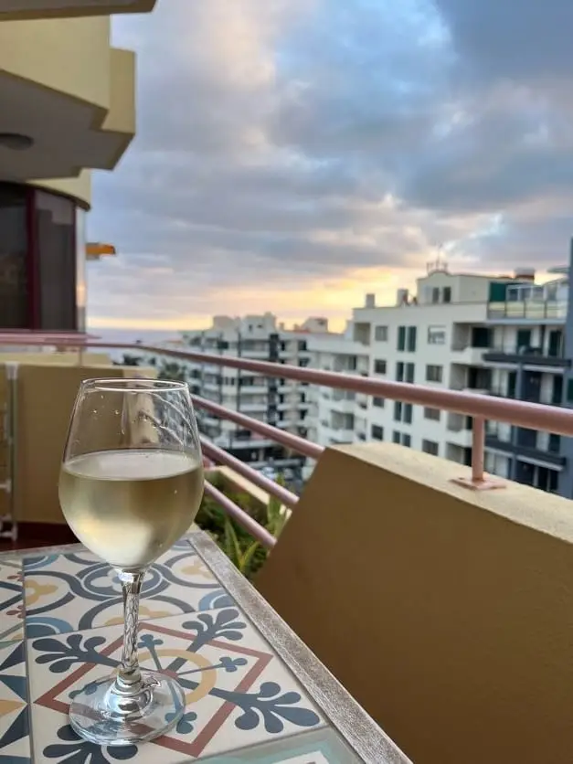 Weinglas auf einem gekachelten Tisch auf dem Balkon, dahinter die Häuser von Funchal in der Abenddämmerung