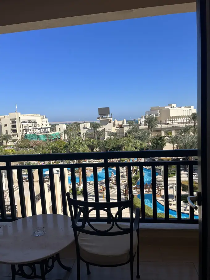 Blick vom Balkon mit Tisch und Stuhl über Palmen und Pools zu weiteren Gebäuden der Anlage, dahinter ein schmaler Streifen Meer