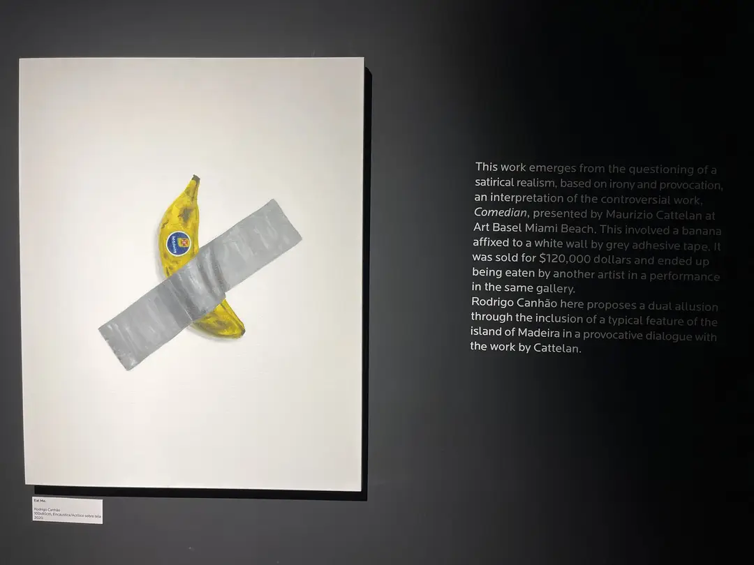 Eine mit grauem Klebeband an eine weiße Wand geklebte Banane, daneben eine erklärende Texttafel