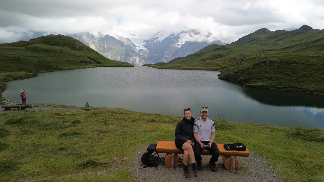 Drohnenfoto: Max und Rebecca sitzen auf einer Holzbank vor dem Bachalpsee, dahinter grüne Hänge und Berge mit Gletscher in den Wolken