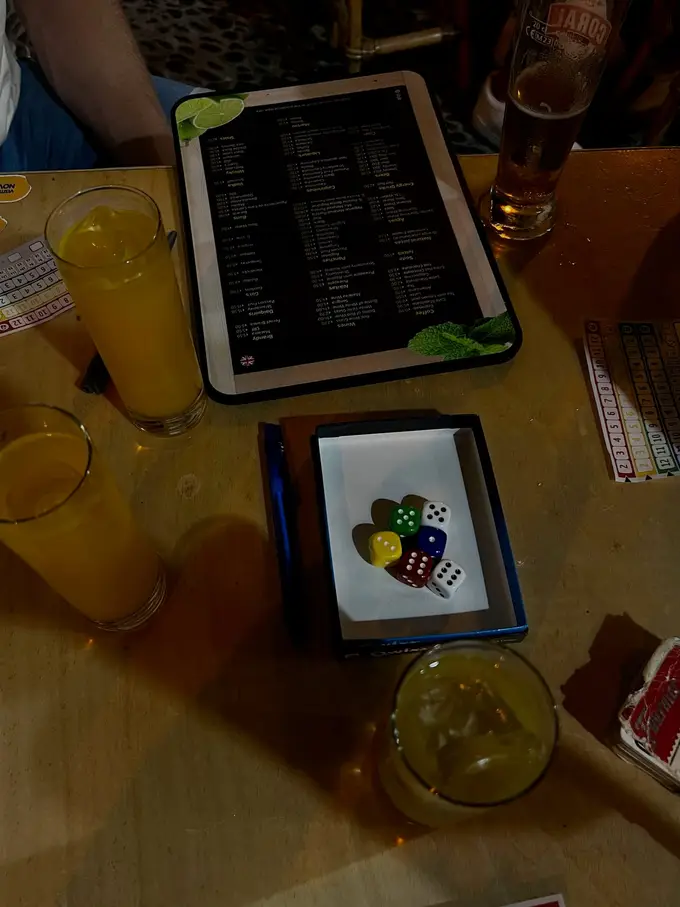 Zwei orangefarbene Cocktails und ein Bier auf einem Tisch, dazu eine Speisekarte und ein Würfelbecher