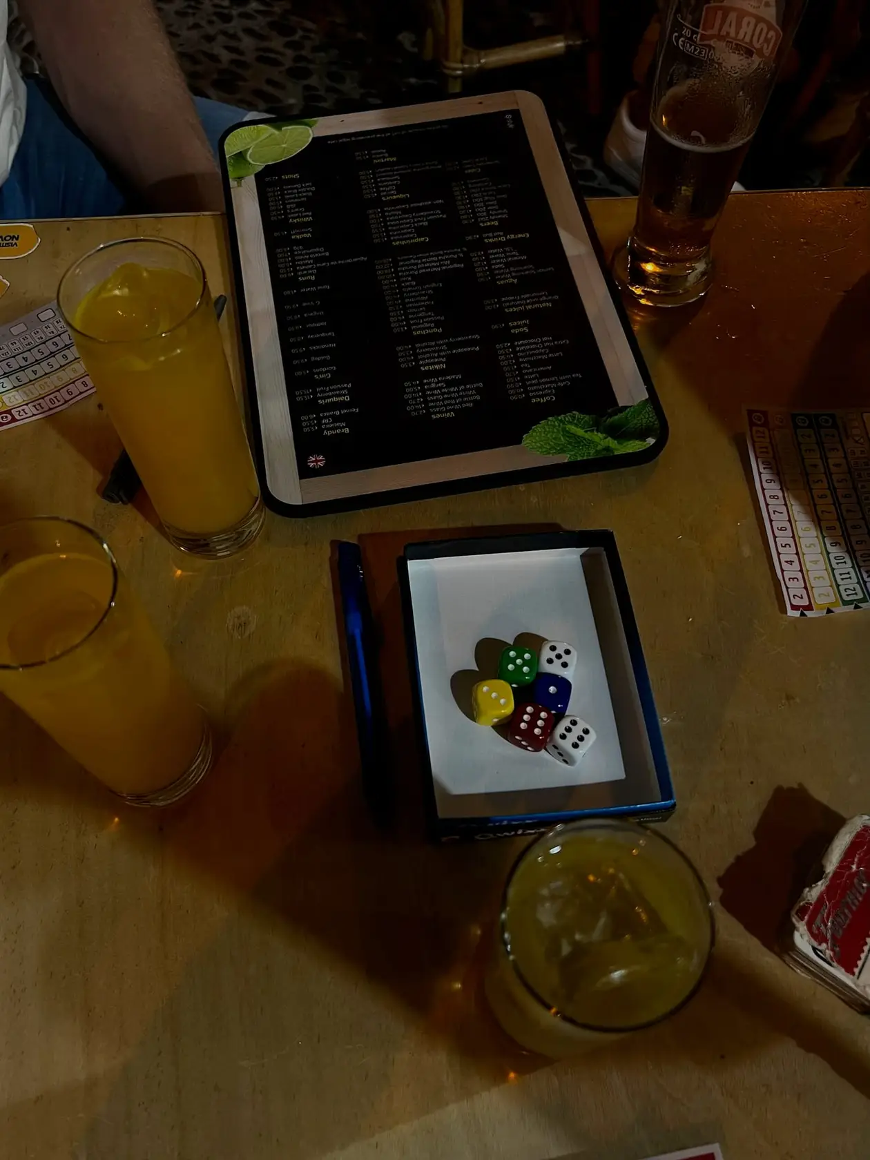 Zwei orangefarbene Cocktails und ein Bier auf einem Tisch, dazu eine Speisekarte und ein Würfelbecher