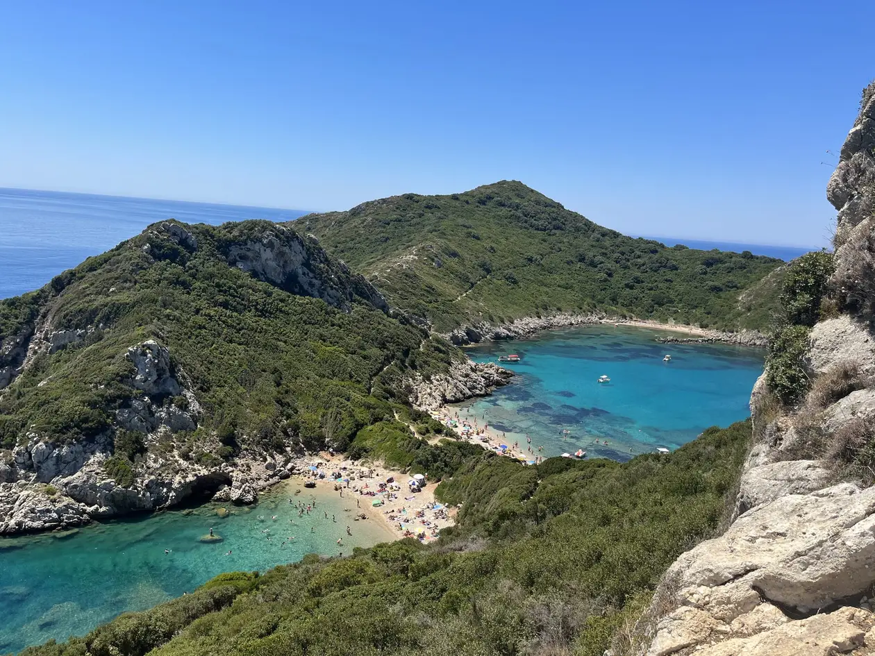 Zwei türkise Buchten mit hellen Stränden, getrennt von einer schmalen bewachsenen Landzunge, rechts ein Felsen im Vordergrund