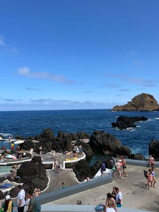 Mehrere Personen liegen und stehen an den Lavabecken, im Hintergrund eine felsige Landzunge im Meer