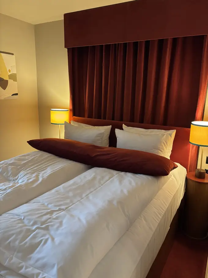 Doppelbett mit weißer Bettwäsche und einem rotbraunen Kissen vor einem dunkelroten Samtvorhang, links und rechts Nachttischlampen mit gelben Schirmen