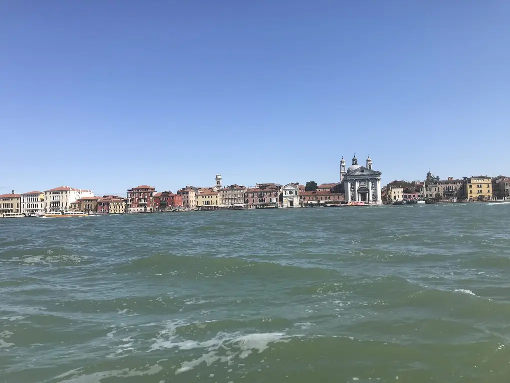 Blick vom Boot über den breiten Kanal auf eine lange Häuserzeile mit einer Kirche mit weißer Fassade
