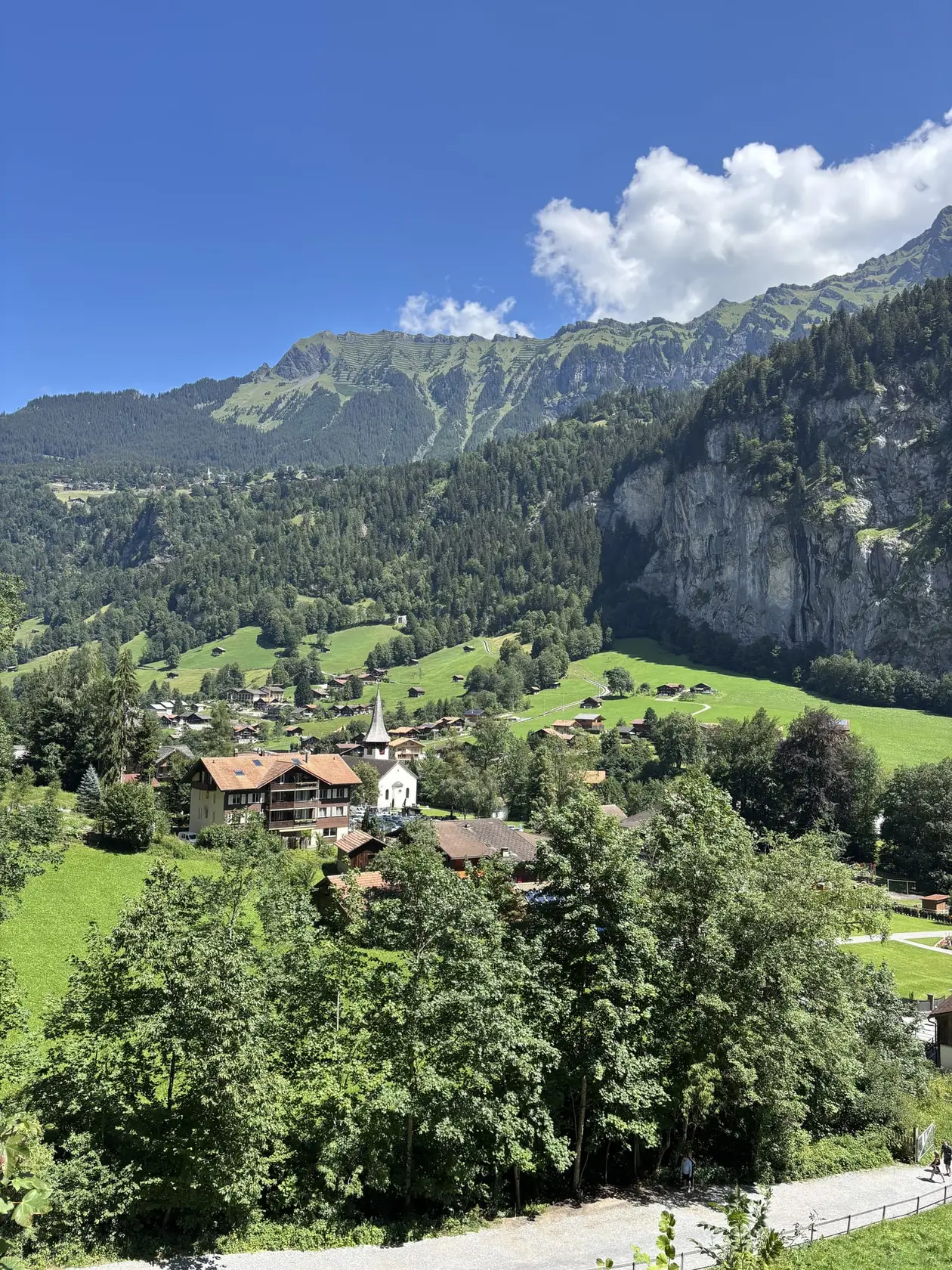 Blick von oben auf ein Dorf mit Kirche im grünen Talboden, rechts eine steile Felswand, dahinter bewaldete Hänge und ein Grat