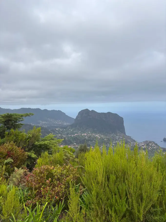 Aussicht von einem Pass über bewachsene Hänge hinunter auf ein Dorf an der Küste und einen markanten Felsen im Meer