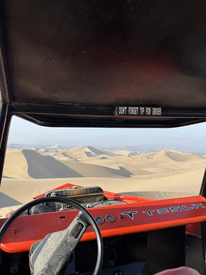 Blick vom Beifahrersitz über Lenkrad und rotes Armaturenbrett auf die Dünen, am Dach ein Aufkleber, der ans Trinkgeld für den Fahrer erinnert