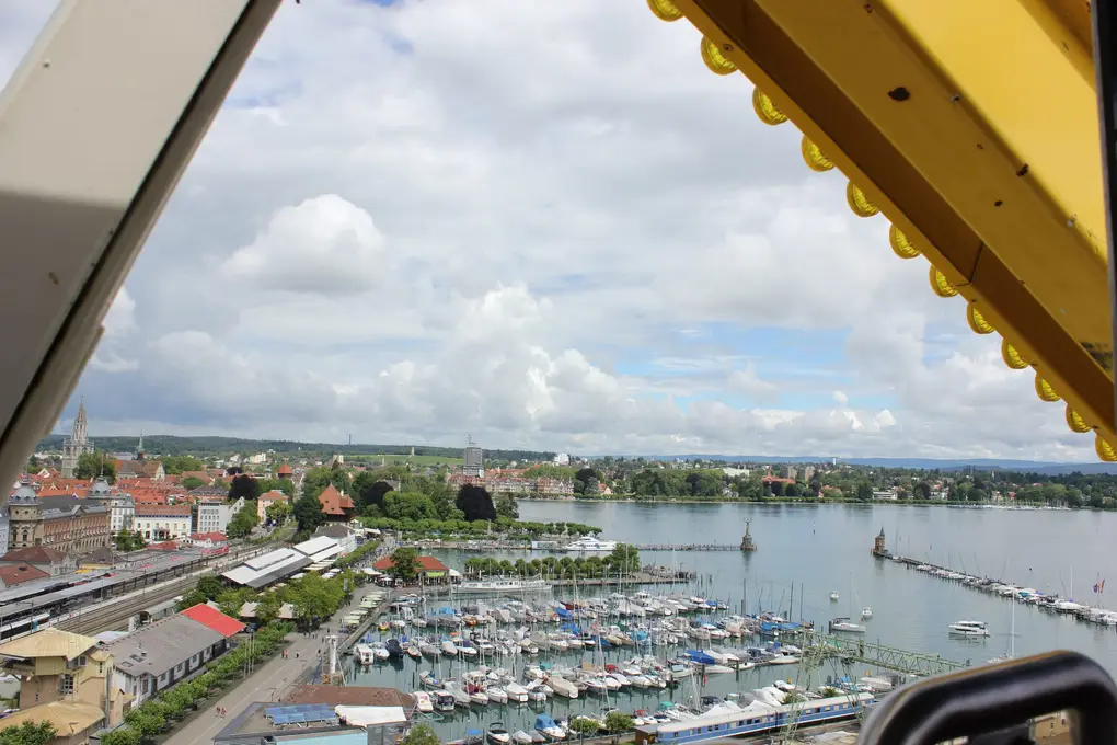 Blick aus der Gondel über den Hafen von Konstanz mit vielen Segelbooten, links die Altstadt mit dem Münsterturm, am Rand gelbe Teile des Riesenrads