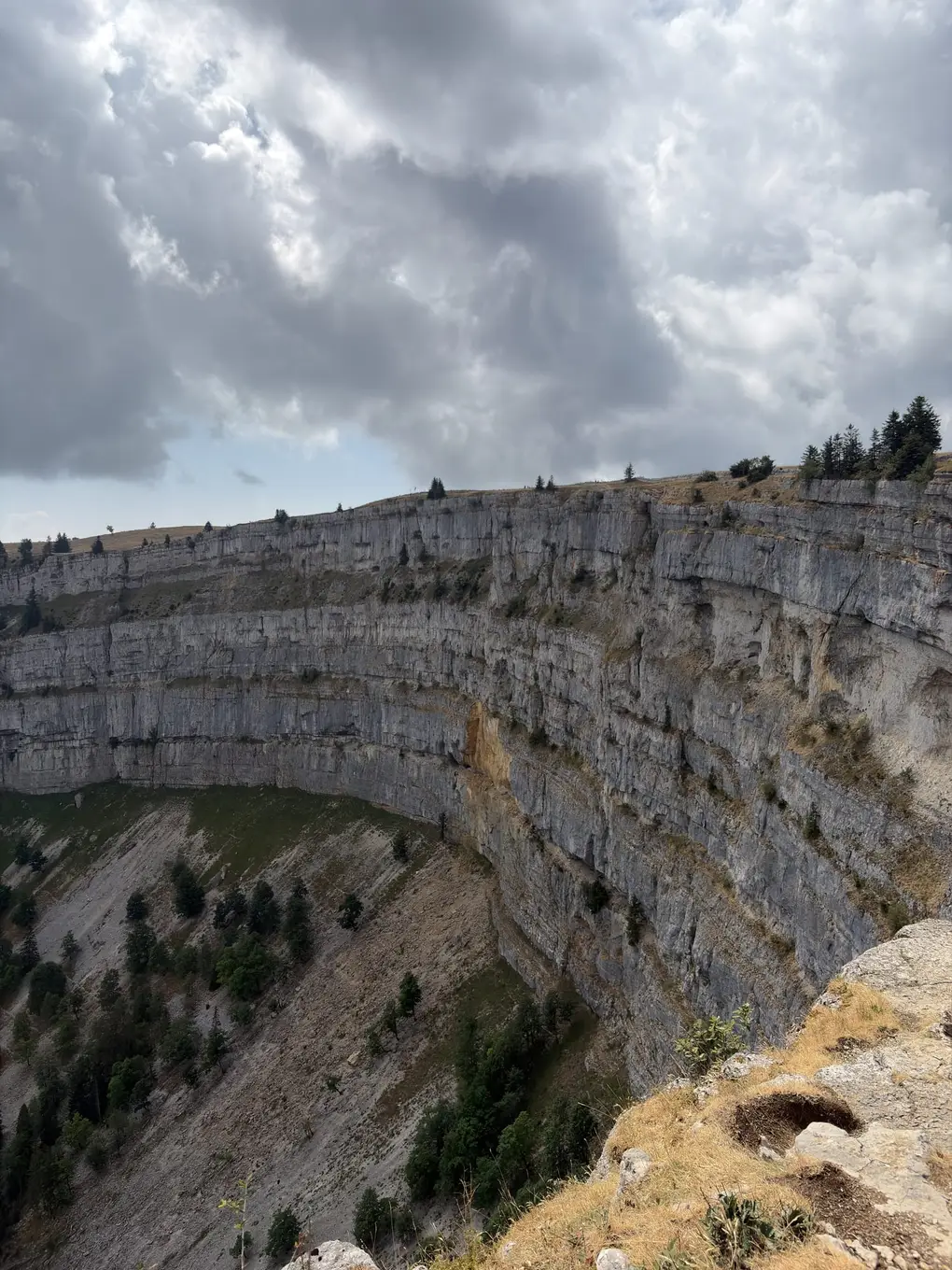 Blick von einer grasigen Felskante entlang der gebogenen Wand in den Kessel hinunter, darüber dunkle Wolken
