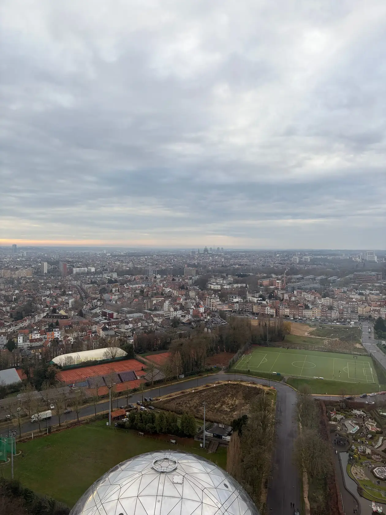 Blick von oben aus dem Atomium über ein weites Häusermeer bis zum Horizont, unten eine silberne Kugel, Sportplätze und Tennisplätze