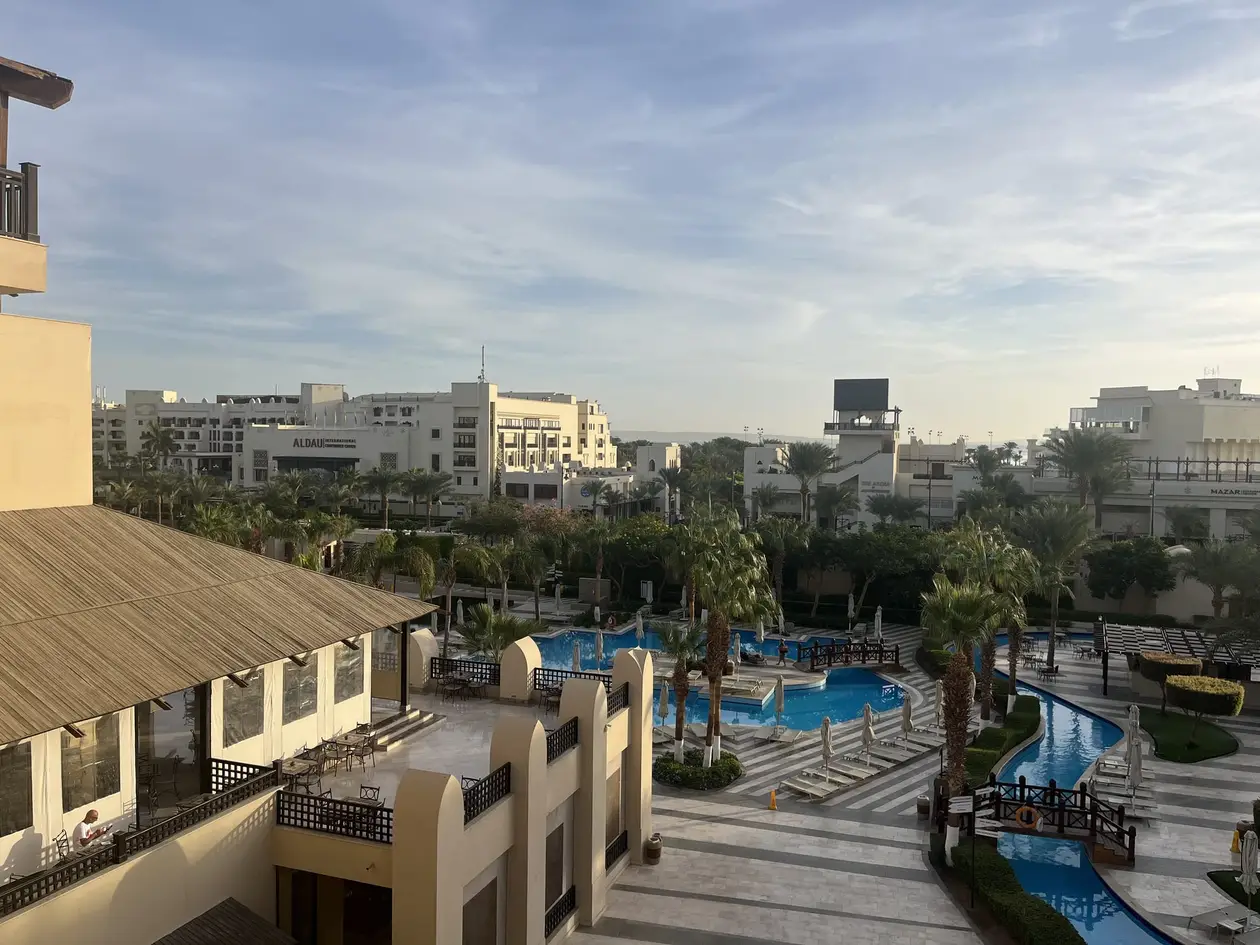 Blick von oben über die Hotelanlage am Morgen, Poolbecken mit Holzbrücken, leere Liegen, Palmen und helle Gebäude