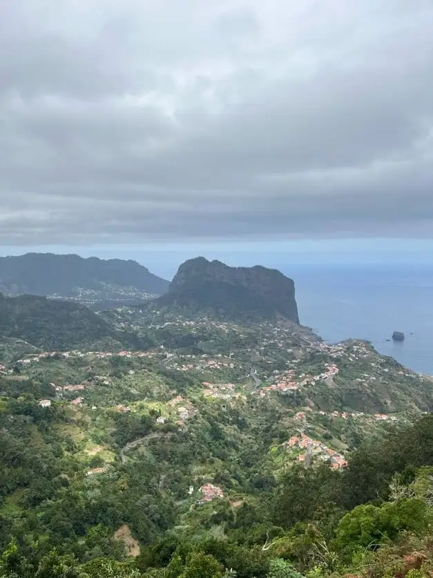 Blick von einem Aussichtspunkt über bewachsene Hänge auf einen schroffen Felsen an der Küste, dahinter das Meer