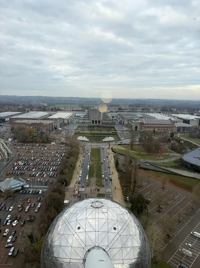Blick von oben aus dem Atomium auf eine silberne Kugel, Parkplätze, eine Allee und die Messehallen am Heysel