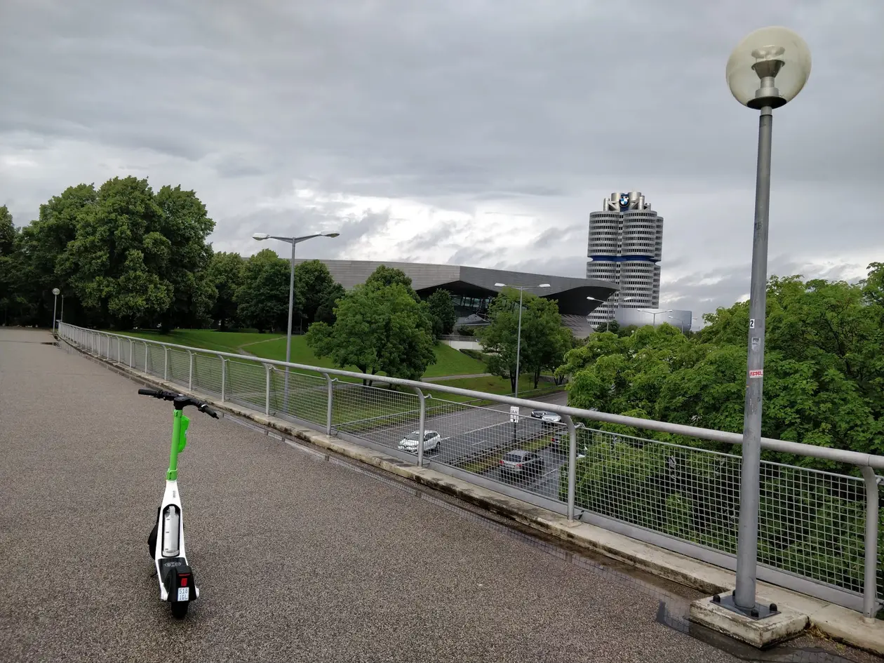 Blick von einer Brücke über eine Straße auf die geschwungene BMW Welt und den runden BMW-Turm, vorne ein abgestellter E-Roller