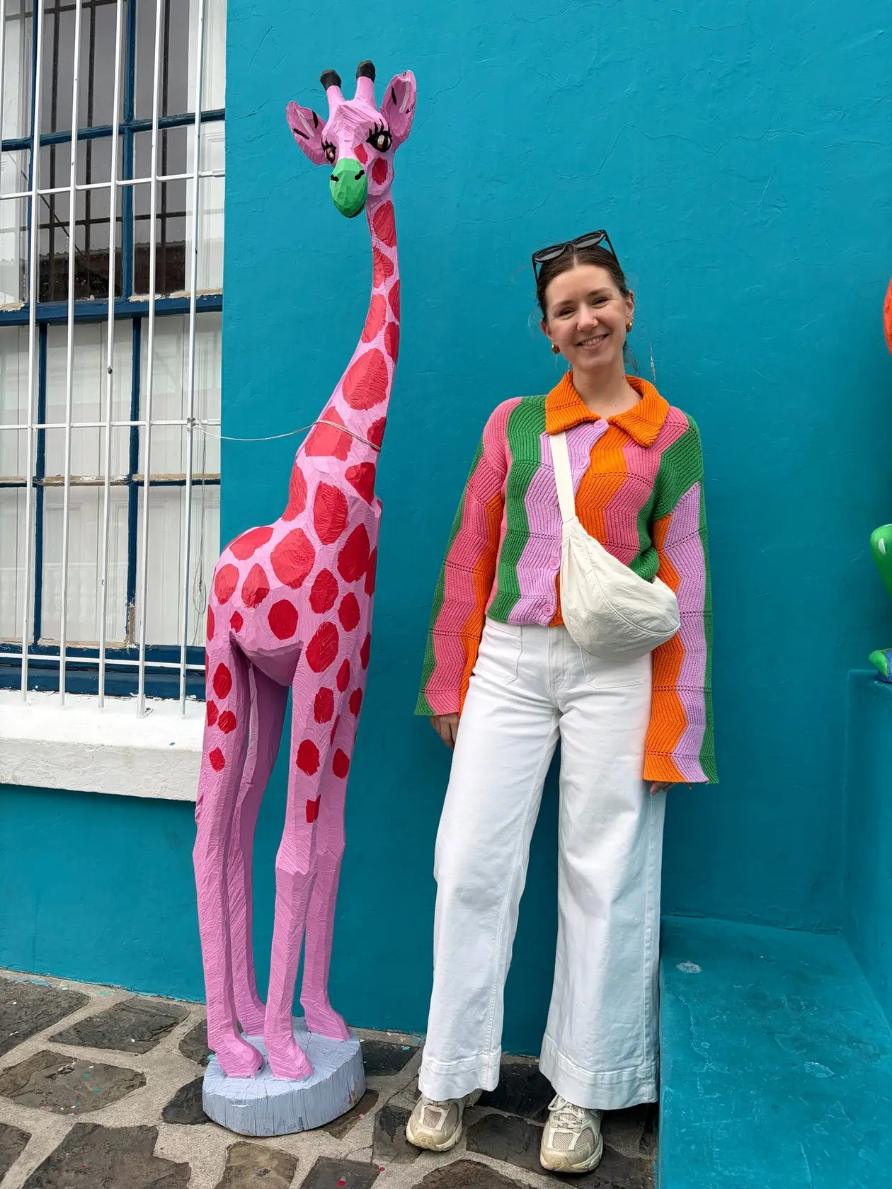 Rebecca steht lächelnd neben einer pinkfarbenen Giraffenskulptur vor einer türkisfarbenen Hausfassade mit der Aufschrift „Art Gallery“