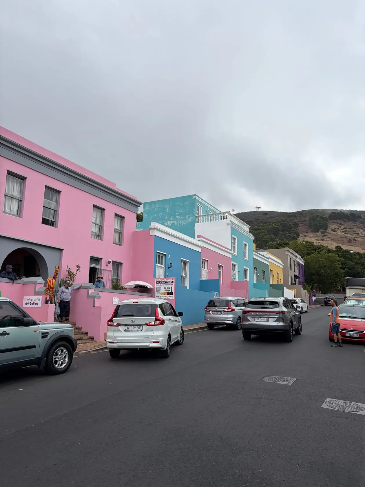 Straße mit bunt gestrichenen Häusern in Rosa, Blau und Türkis am Hang von Bo-Kaap, geparkte Autos am Straßenrand