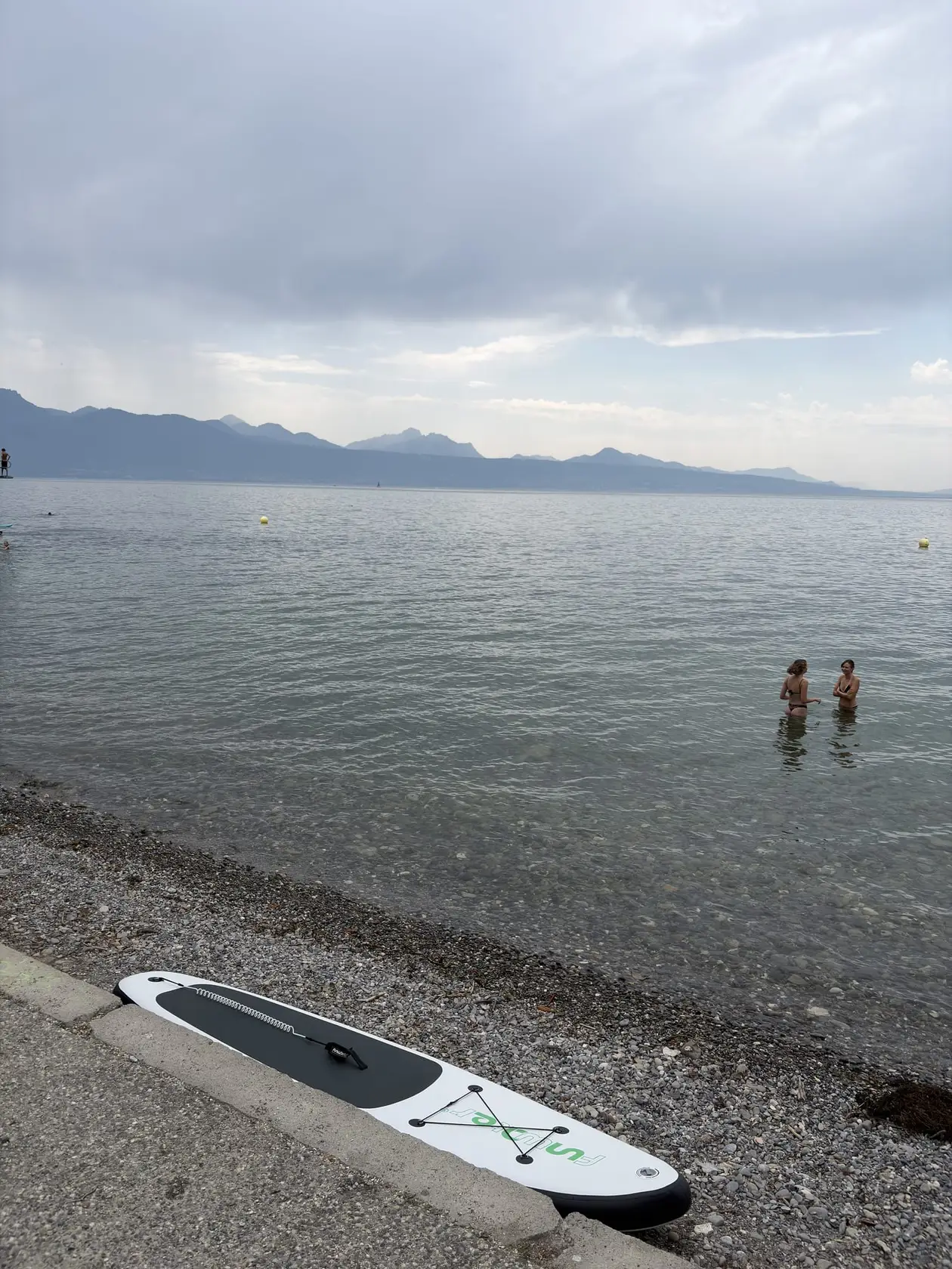 Ein Stand-up-Paddle-Board liegt am Kiesufer des Genfersees, im Wasser zwei Badende, in der Ferne Berge unter Wolken