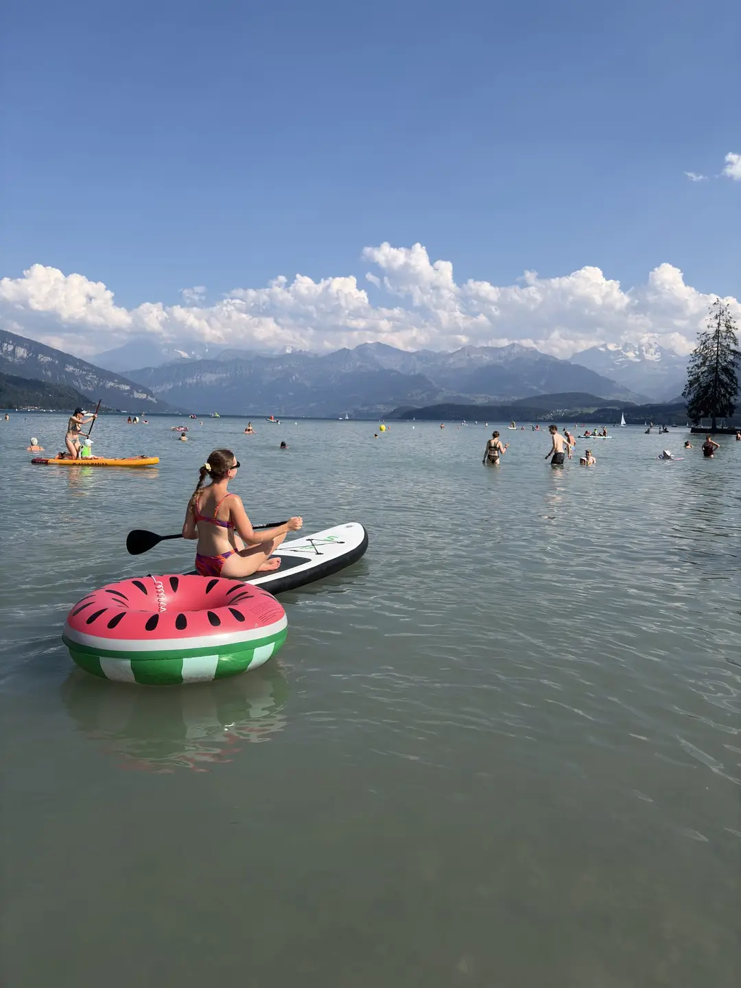 Rebecca sitzt paddelnd auf einem Stand-up-Paddle-Board, davor ein Schwimmring im Wassermelonen-Look, dahinter Badende und Berge am Thunersee
