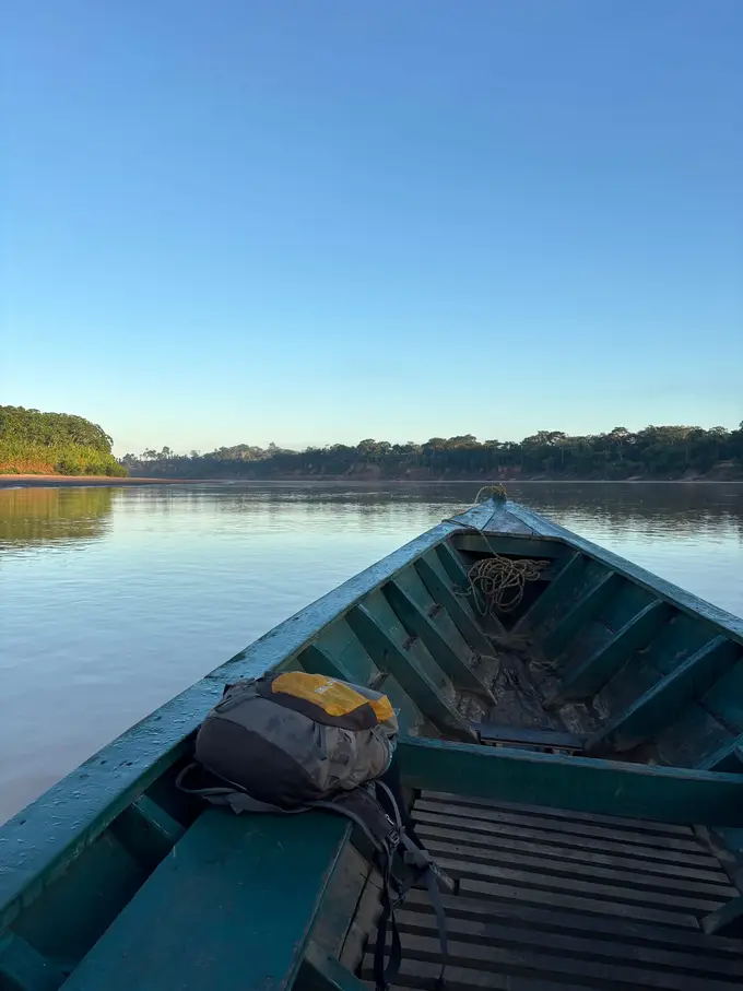 Blick über den Bug eines Holzboots mit Rucksack auf den glatten Fluss im Morgenlicht, am Ufer Wald