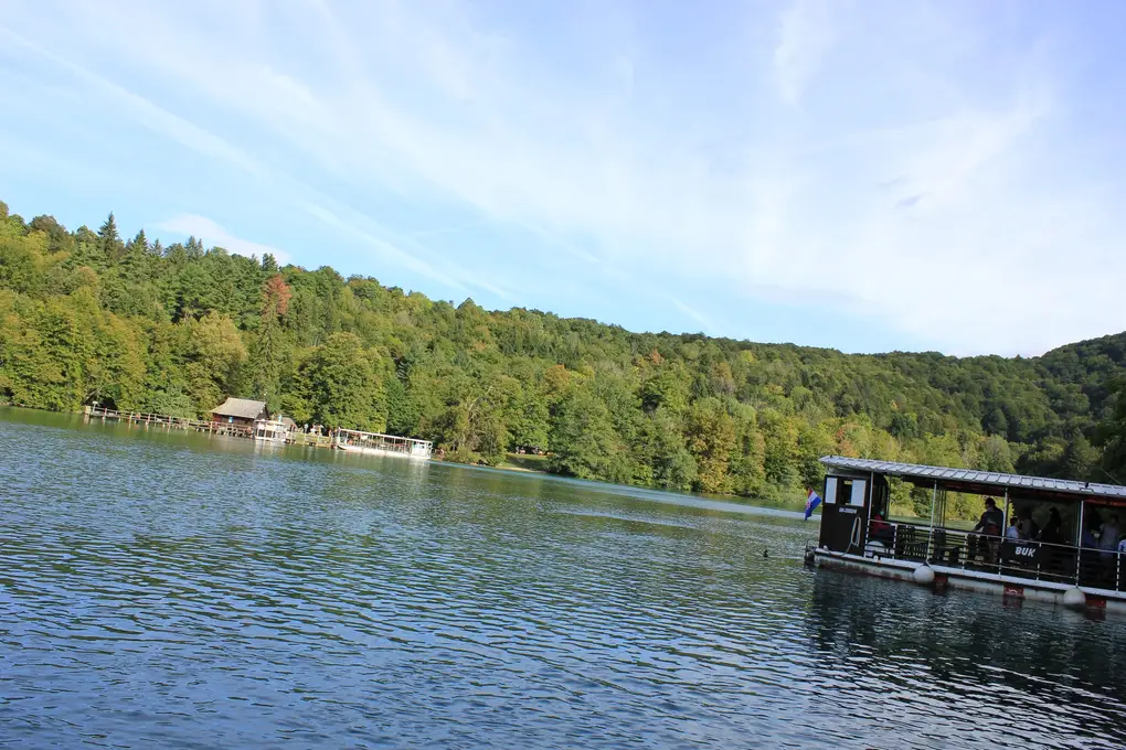 Ein Elektroboot mit Fahrgästen und kroatischer Flagge auf einem See, am anderen Ufer ein Anleger mit Holzhaus und Wald